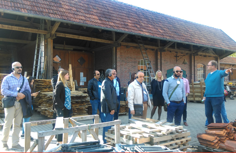 Eine Gruppe steht vor einem bauernhofartigen Gebäude; im Vordergrund sind verschiedene Stapel mit Dachziegeln zu sehen. Ein Mann erklärt etwas und zeigt mit der Hand in eine Richtung.